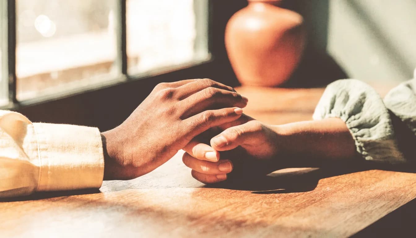 Two people hold hands across a wooden table in warm light, leaning toward each other in a calm, attentive conversation.