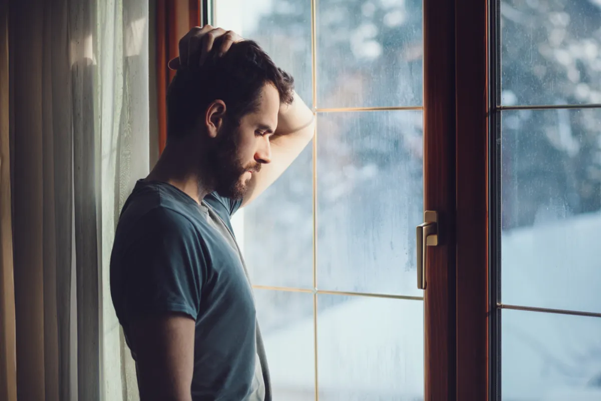A man standing by a window, arms folded, looking thoughtful