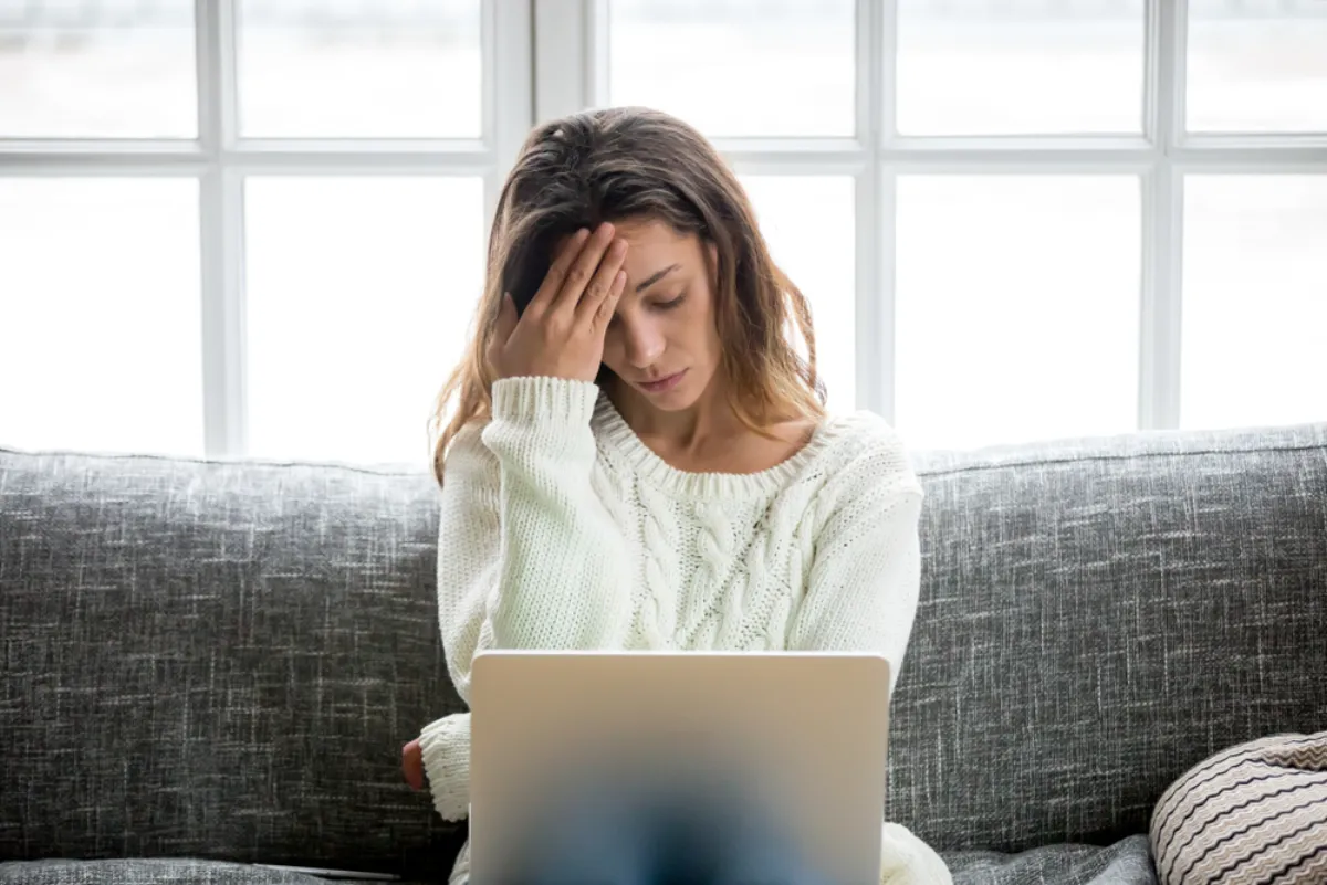 A woman sitting on a sofa with a laptop, looking troubled