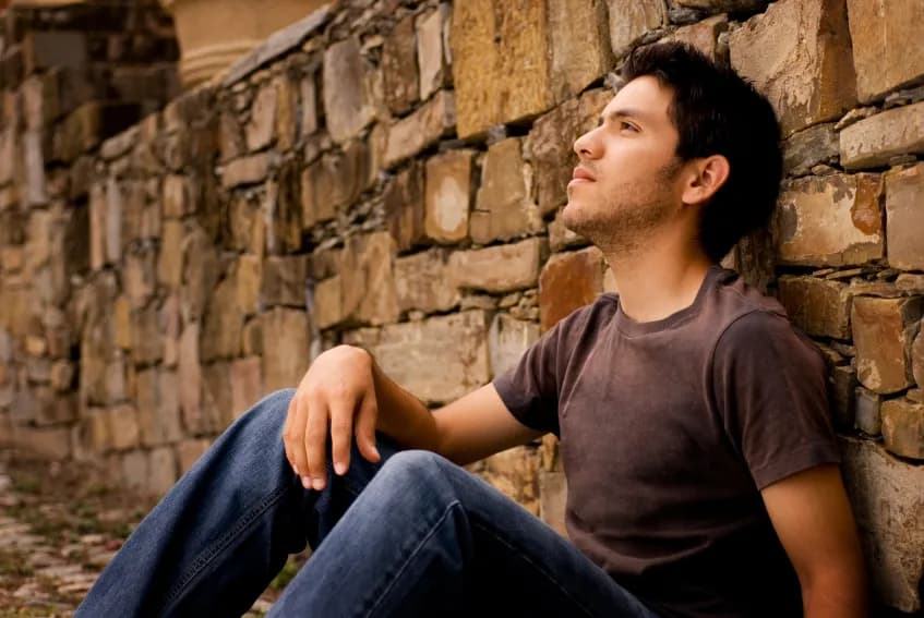 A young man sitting against a stone wall, looking up thoughtfully