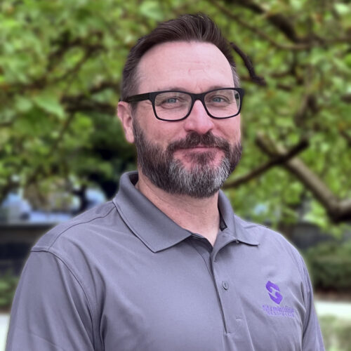Christopher Knox, Clinical Mental Health Counselor (CMHC) at Connections Counseling, smiling outdoors in a gray polo with the Streamline logo. Chris specializes in working with youth, families, and adults, providing individual, family, and group therapy.